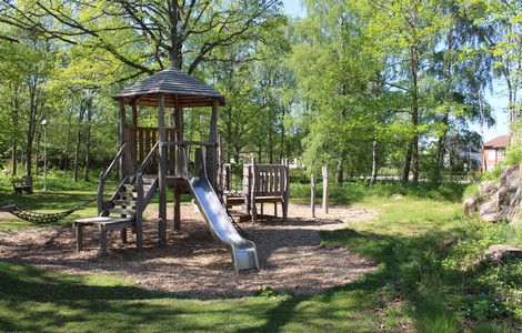 En lekpark i ett grönområde med grönskande träd. Mitt på bilden finns ett lektorn byggt av trä med tillhörande rutschkana.