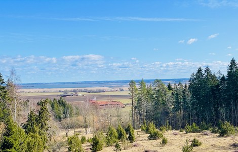Landskapsfoto över Ombergslidens naturreservat. På bilden ser man en höjd med skog på höger sida. Längre fram ser man ett öppet odlingslandskap, en sjö och en klosterbyggnad.