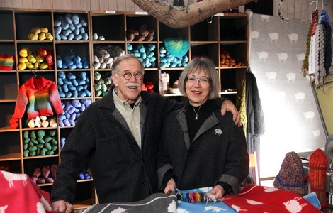 Ett porträttfoto av ägarna till Östergötlands ullspinneri. Bilden föreställer en man & en kvinna som står och poserar i en textilbutik. I bakgrunden ser man en hylla med flera fack som innehåller garn i olika färger. 