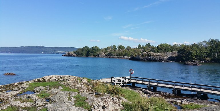 En vy bild över Stora Lunds badplats. På bilden ser man en klippa och precis nedanför sitter en brygga. På bryggan sitter en livboj. Bakom ser man vattnet och i bakgrunden skymtar land. 