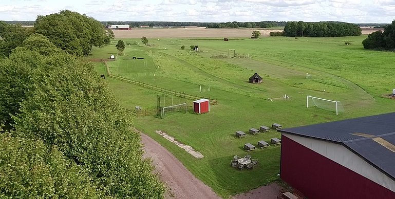 Ett flygfoto över Sjöstorps fotbollsgolf. På bilden ser man en stor gräsplan med olika hinder. I högra hörnet står ett rött magasin och till vänster reser sig stora gröna buskage. 