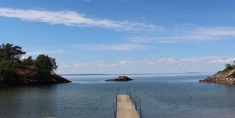 Ett foto över Hästholmens badplats. På bilden ser man en lång brygga av trä som är placerad i en vik. På höger och vänster sida ser man varsin udde och i mitten skymtar en liten stenhäll från vattnet. 