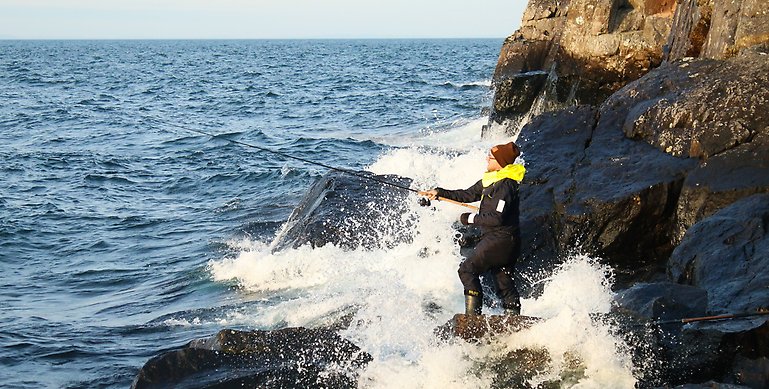En man som står och fiskar på ett par klippor när vattnet slår mot land. Mannen har ett fiskespö pch har på sig en svart fritidsjacka, gul hoodietröja och en röd mössa.